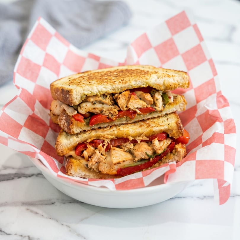 Grilled chicken sandwich with red peppers and onions, served in a red and white checkered paper.