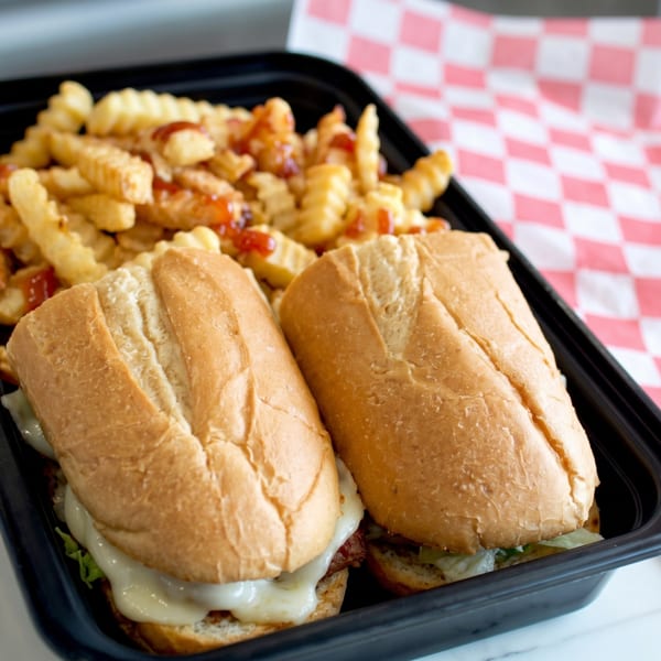 Two cheesesteak sandwiches and crinkle-cut fries with ketchup in a black to-go container.