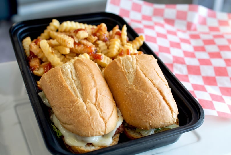 Two cheesesteak sandwiches and crinkle-cut fries with ketchup in a black to-go container.