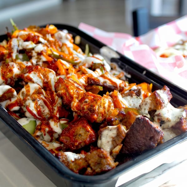 Close-up of a black takeout container filled with saucy chicken and vegetables, with fries in the background.