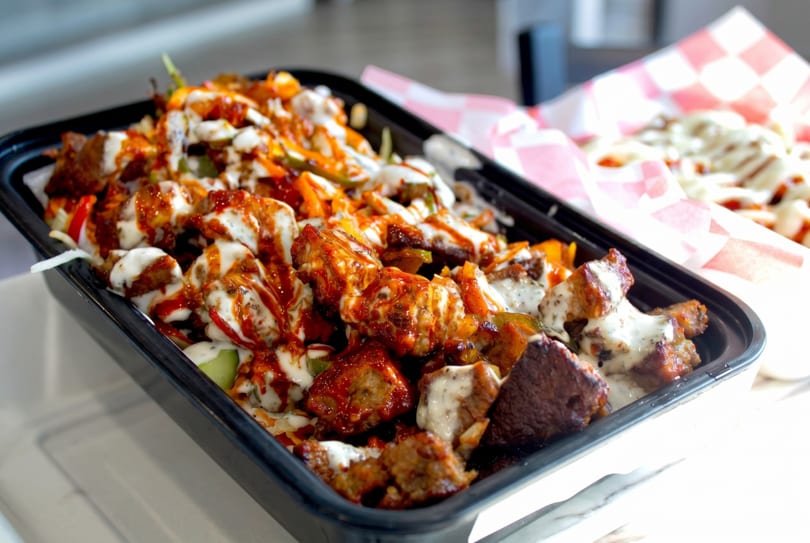 Close-up of a black takeout container filled with saucy chicken and vegetables, with fries in the background.