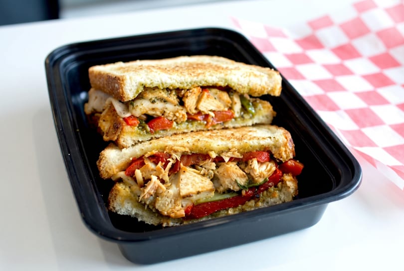 Two halves of a grilled chicken and roasted red pepper sandwich in a black to-go container.