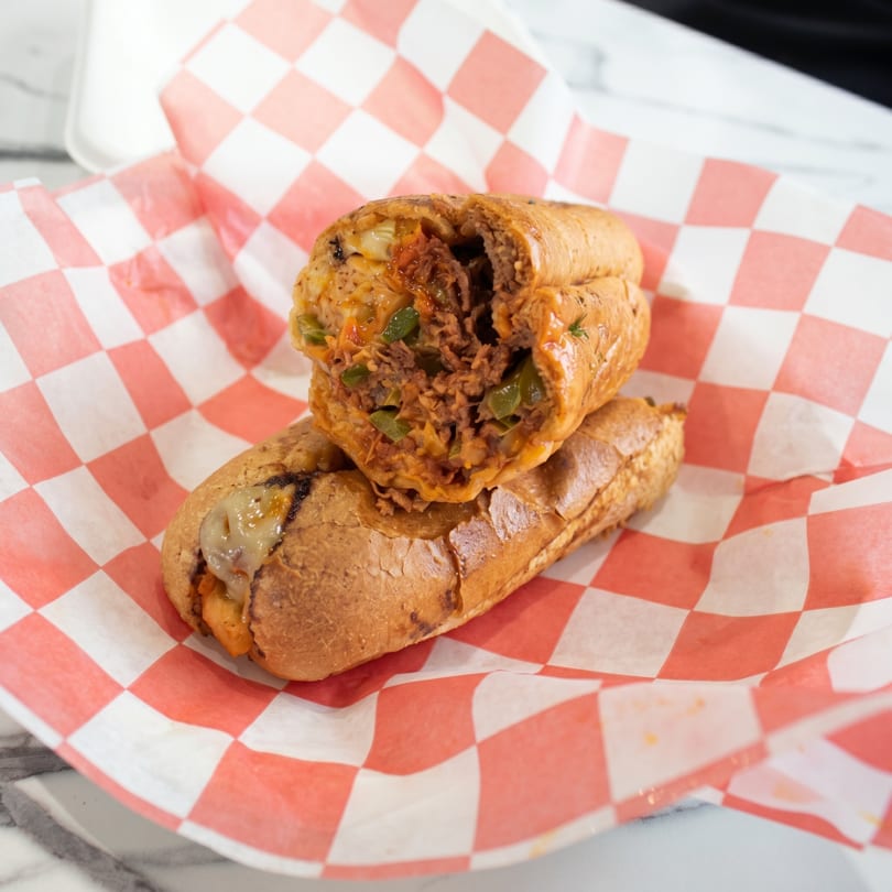 Cheesesteak sandwich with melted cheese and green peppers, served on red and white checkered paper.